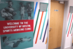Door to appointment room at the indoor sports centre. The sign on the wall says 'welcome to the university of bristol sports medicine clinic'. There are pencil sketch images of students playing sports in the background of the wall.